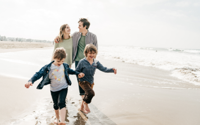 A family of four is walking together. The mom and dad are behind the little boy and girl walking in the sand. alongside the beach.