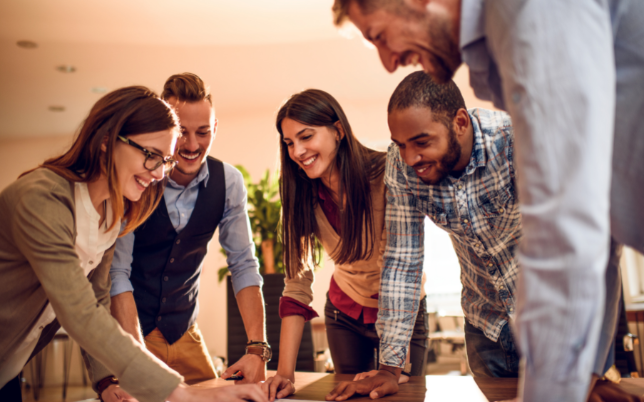 Five employees have surrounded a table smiling and talking.